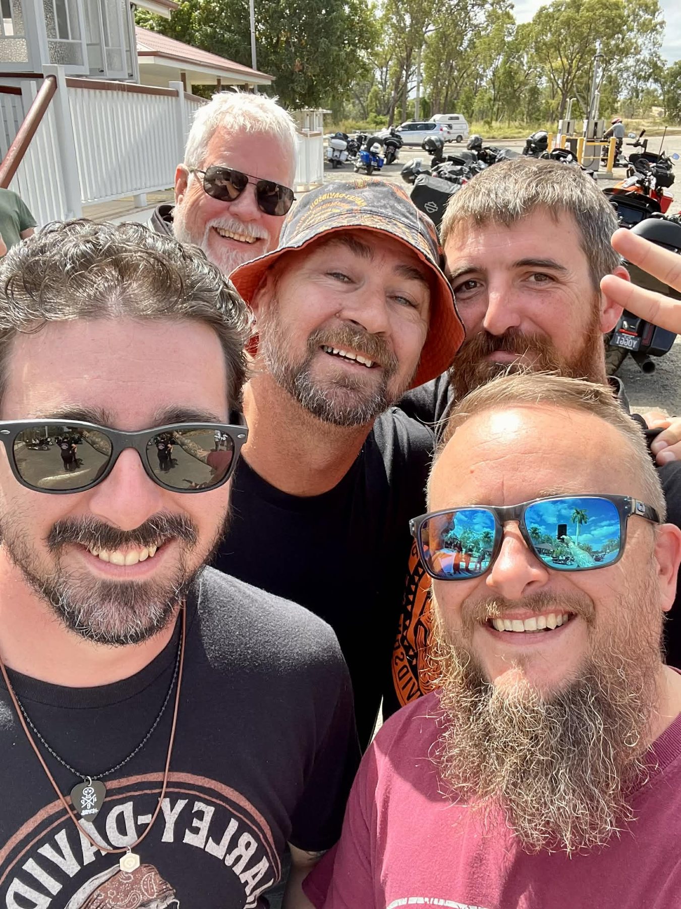 Five rally riders smiling for a group selfie with motorcycles behind them.